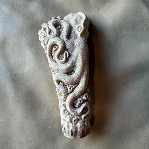 Könnte beinhalten: Ein cremefarbenes, zylindrisches Objekt mit einer komplexen Hochreliefschnitzerei eines Oktopus. Die Tentakel des Oktopus winden sich um die Oberfläche und zeigen detaillierte Saugnäpfe sowie strukturierte Hautmuster über die gesamte Länge.
