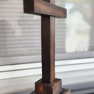May include: A dark brown wooden cross featuring a vertical post and a horizontal crossbar, mounted on a square base. The wood displays a visible grain pattern with varying shades of brown, and the structure has a smooth, matte finish throughout.