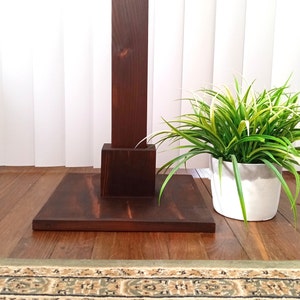 May include: A dark brown wooden plant stand with a square base and a single upright post. The stand is on a wooden floor with a patterned rug in the foreground. A potted plant with green and white leaves is in the background.