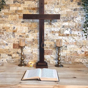 May include: A dark brown wooden cross stands upright on a base, flanked by two metallic candle holders with glass inserts. An open book with white pages lies flat in the foreground, and a small, colorful patterned bowl sits near the cross.
