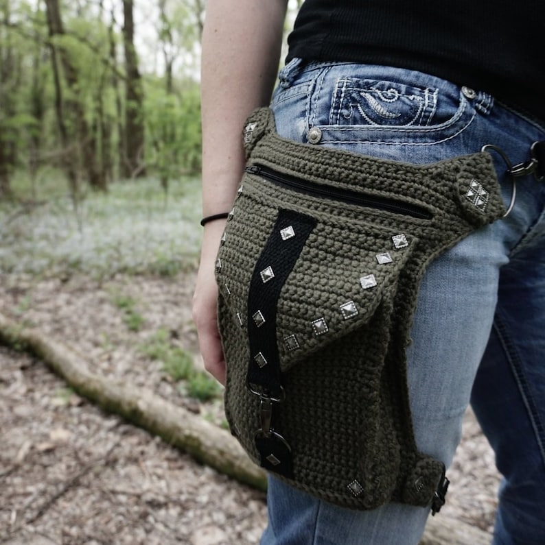 May include: A green crocheted fanny pack with silver studs and a black strap. The pack is worn on the hip of a person wearing blue jeans.