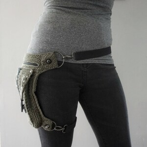 May include: A green crocheted thigh holster with a black belt and silver hardware. The holster is worn on the left leg and has a small pocket on the front.
