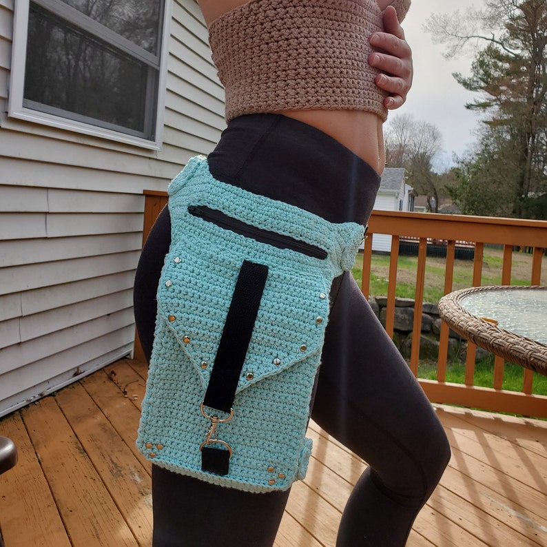 May include: A light blue crocheted hip bag with a black strap and silver studs. The bag has a zippered pocket and a flap with a snap closure.