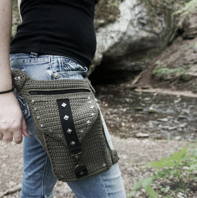 May include: A green crocheted hip bag with silver studs and a black strap. The bag is worn over blue jeans.