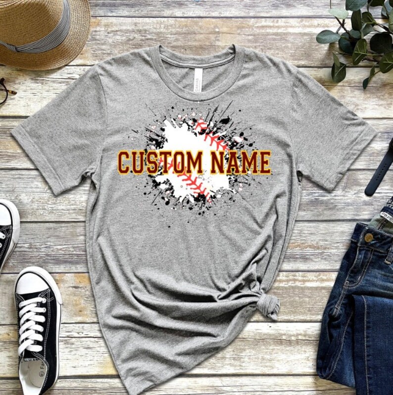 May include: Gray t-shirt with a white baseball graphic and black splattered paint. The text "CUSTOM NAME" is in gold letters.