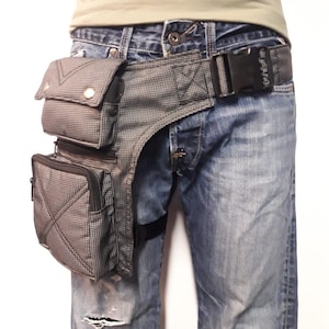 Puede incluir: Una bolsa de muslo a cuadros en negro y gris con m&uacute;ltiples bolsillos y un cintur&oacute;n negro ajustable. La bolsa se lleva en la pierna derecha de una persona que lleva pantalones vaqueros azules.