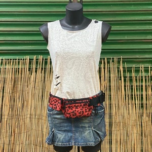 Puede incluir: Una riñonera roja y negra con estampado de leopardo con un cinturón y hebilla negros. La riñonera se lleva sobre un top blanco sin mangas y una falda vaquera azul.