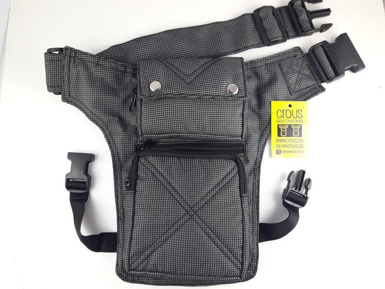 Puede incluir: Una bolsa de muslo a cuadros en negro y gris con m&uacute;ltiples bolsillos y correas ajustables. La bolsa tiene un bolsillo con cremallera en la parte delantera y un bolsillo con cremallera m&aacute;s peque&ntilde;o en el lateral. La bolsa tiene una hebilla negra en la correa. La bolsa tiene una etiqueta amarilla con el texto "Crous Eat Color Live" y una direcci&oacute;n de sitio web.