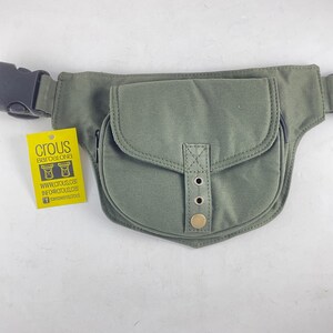 Op de afbeelding: Olijfgroene heuptas met een gebogen klepzak en verstelbare riem. De tas heeft een zwarte gesp en een klein label met de tekst "CROUS BARCELONA". De tas heeft een ritsvak en metalen klinknagels.