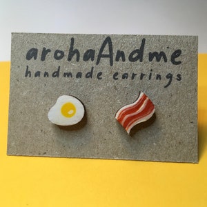 May include: A pair of handmade earrings featuring a fried egg and a piece of bacon. The earrings are made of wood and are painted to look like real food. The earrings are displayed on a brown card with the text "arohaAndme handmade earrings".