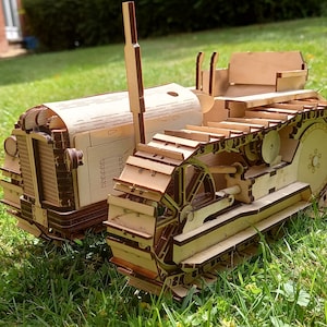 Pode incluir: Um modelo de madeira de um trator vintage em tons de marrom e bege. O modelo apresenta um design detalhado com engrenagens e esteiras visíveis. O trator está sentado em um leito de grama verde.