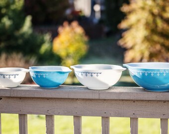 May include: Three blue and white mixing bowls of different sizes, lined up on a wooden railing. The bowls have a white background with a blue and white geometric pattern.