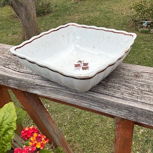 May include: A white, square ceramic bowl with a scalloped rim and a brown border. The bowl features a brown floral design in the center. The bowl is sitting on a wooden railing with red flowers in the foreground.