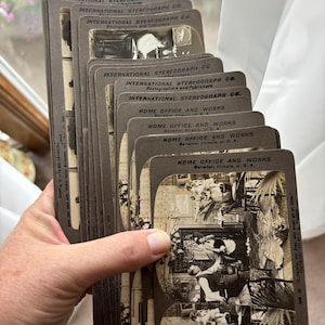 May include: A collection of vintage stereograph cards, held in a hand. The cards are rectangular with rounded corners and feature black and white images. Text on the cards includes "INTERNATIONAL STEREOGRAPH CO." and "HOME OFFICE AND WORKS".