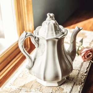 May include: White porcelain teapot with a decorative lid and a fluted design. The teapot is sitting on a wooden surface with a distressed white paint finish.
