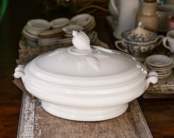 May include: White porcelain serving dish with a lid. The lid has a decorative knob in the shape of a fruit. The dish is on a wooden surface.
