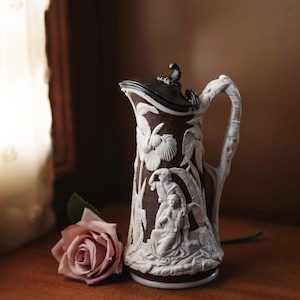 May include: A brown and white ceramic pitcher with a lid and a handle. The pitcher is decorated with a relief design of figures and foliage. A pink rose is in the foreground.