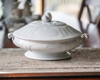 May include: A white ceramic serving dish with a lid, featuring a decorative floral design. The dish has a pedestal base and handles on either side. The lid is topped with a small, decorative knob. The dish is sitting on a weathered wooden surface.