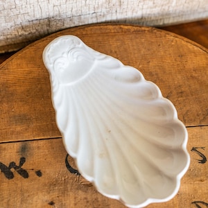 May include: White ceramic dish shaped like a seashell with a decorative design on the top. The dish is sitting on a wooden surface.