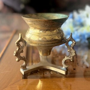 May include: A vintage brass incense burner with a wide, fluted bowl and ornate handles. The burner has a tripod base and a tarnished, aged appearance, suggesting it is an antique. The design is likely for burning incense or holding potpourri.