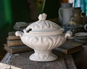 May include: White ceramic serving dish with a lid and ornate detailing. The dish is on a wooden table with books stacked behind it.