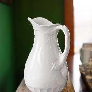 May include: A white ceramic pitcher with a decorative handle and floral design. The pitcher is sitting on a wooden surface.