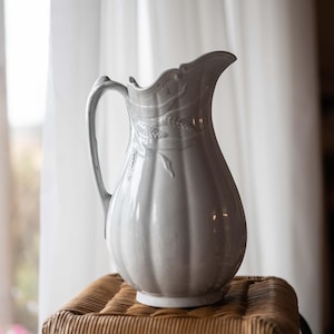 May include: A white ceramic pitcher with a floral design sits on a woven wicker stool. The pitcher has a wide mouth and a handle.