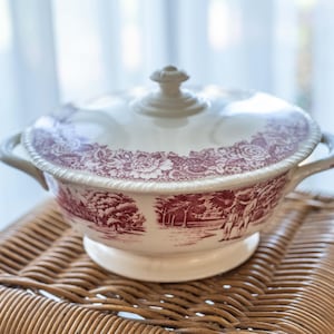 May include: A white ceramic serving dish with a red floral pattern and a lid. The dish is sitting on a woven wicker surface.
