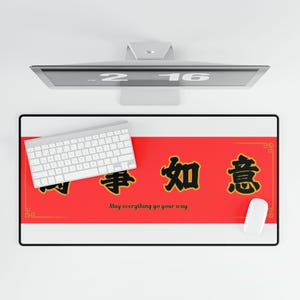Puede incluir: Alfombrilla de escritorio rectangular con un panel central rojo con caracteres chinos y la frase en inglés "May everything go your way". Un teclado y un ratón blancos están sobre la alfombrilla. La alfombrilla está enmarcada en negro.