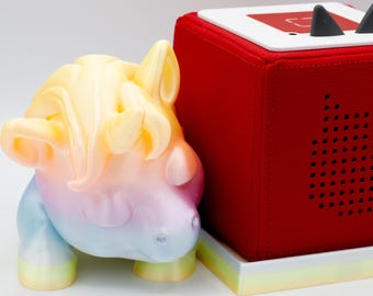 May include: A red cube-shaped audio player with black horns and a white top, next to a colourful unicorn figurine. The unicorn is a mix of yellow, pink, and blue hues. The audio player sits on a rectangular base.