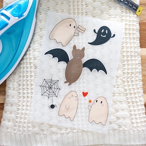 Può includere: Un foglio di carta transfer termoadesiva con una varietà di disegni a tema Halloween, tra cui fantasmi, pipistrelli e una ragnatela. I disegni sono stampati in nero, marrone e bianco su uno sfondo trasparente. La carta è appoggiata su un tessuto bianco a maglia con un ferro da stiro blu e un metro a nastro.