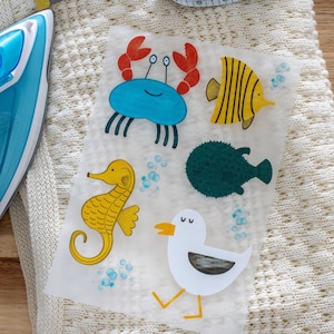Könnte beinhalten: Bügelbild mit fünf bunten Meeresbewohner-Motiven: ein blauer Krebs, ein gelber Fisch, ein grüner Kugelfisch, ein gelbes Seepferdchen und eine weiße Möwe.