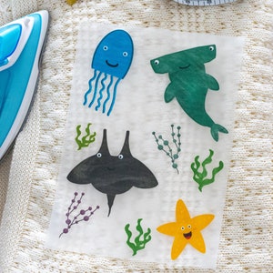 May include: A sheet of iron-on fabric transfers featuring sea creatures: a blue jellyfish, a green hammerhead shark, a black stingray, and a yellow starfish. A measuring tape and iron are also visible. The background is a cream-colored knitted fabric.