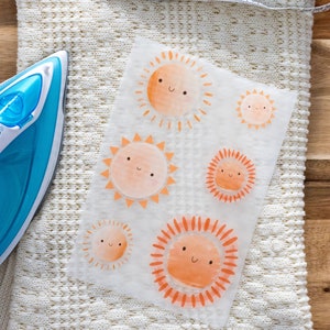 Può includere: Un foglio di carta transfer per stirare con cinque disegni di sole arancioni. Ogni sole ha un viso sorridente e un bagliore arancione radiante. La carta transfer è appoggiata su una coperta di maglia bianca con un metro a nastro e un ferro da stiro blu.