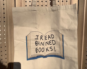 Puede incluir: Un bolso tote de lona beige con asas largas. La parte delantera del bolso presenta un gráfico de un libro abierto con el texto "I READ BANNED BOOKS!" en negro. El contorno del libro es azul.