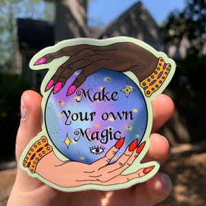 May include: A sticker featuring a crystal ball design with the text "Make your own Magic." The crystal ball is held by two hands with long, painted nails and gold bracelets. The background is a mix of purple, blue, and yellow.