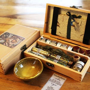 Peut inclure: Deux boîtes en bois clair, l'une fermée et l'autre ouverte, contenant des fioles en verre avec des herbes séchées, un petit bol en céramique et une carte en papier avec le texte Räucherkiste. La boîte fermée porte un autocollant carré illustré.