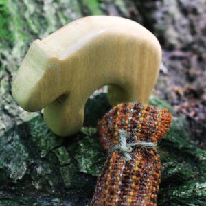 Könnte beinhalten: Eine glatte, hellfarbene Bärenfigur aus Holz mit abgerundeter Form, platziert neben einem kleinen, strukturierten Stoffbeutel mit einem Webmuster aus braunen, orangefarbenen und grauen Fäden, der mit einer einfachen Schnur verschlossen ist.