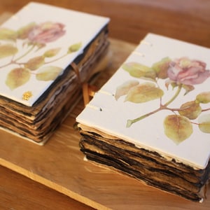 May include: Two rectangular journals featuring white covers with a watercolor illustration of a pink rose and green leaves. The pages have dark, distressed edges, and each book is bound with white thread and includes a brown ribbon with small charms.
