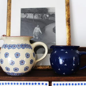 Könnte beinhalten: Ein cremefarbener Keramikkrug mit blauem Blumenmuster und ein kleinerer, dunkelblauer Keramikkrug mit weißem Punktmuster. Beide Krüge haben einen bauchigen Körper, einen geschwungenen Henkel und einen Ausguss am oberen Rand.