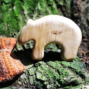 Könnte beinhalten: Eine glatte, helle Holzfigur in Bärenform mit sichtbarer natürlicher Maserung neben einem kleinen, rechteckigen, strukturierten Beutel mit einem Webmuster aus orangefarbenen, braunen und weißen Fäden, der mit einer dünnen Schnur gebunden ist.