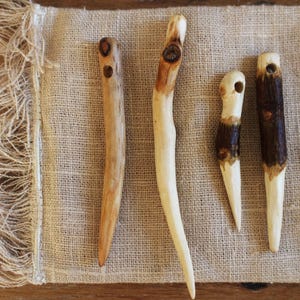 May include: Four hand-carved wooden hair sticks or needles with natural wood grain and knots. Each piece features a tapered, pointed end and a single hole at the top. The colours range from light beige to dark brown, showcasing the organic texture.
