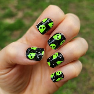 May include: Close-up of a hand with black and green nail wraps featuring alien faces. The nail wraps are shiny and have a black background with green alien faces.
