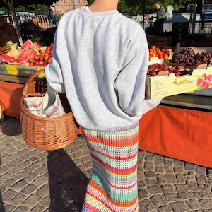 K&ouml;nnte beinhalten: Eine Person tr&auml;gt ein graues Sweatshirt und einen bunten, gestreiften H&auml;kelrock. Die Person h&auml;lt einen Weidenkorb und steht vor einem Obststand.