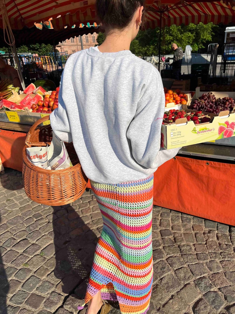 K&ouml;nnte beinhalten: Eine Person tr&auml;gt ein graues Sweatshirt und einen bunten, gestreiften H&auml;kelrock. Die Person h&auml;lt einen Weidenkorb und steht vor einem Obststand.