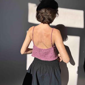 May include: A pink knitted crop top with thin straps. The top is paired with a black skirt. The model is facing away from the camera, with the back and shoulders visible. The lighting creates shadows on the wall.