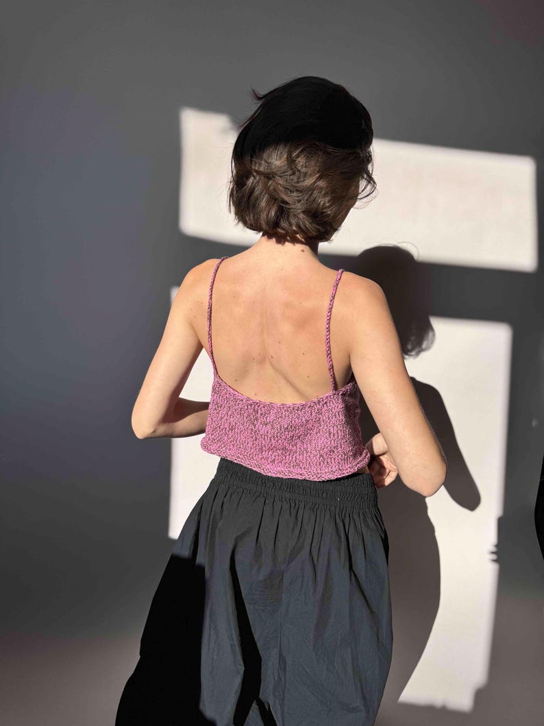 May include: A pink knitted crop top with thin straps. The top is paired with a black skirt. The model is facing away from the camera, with the back and shoulders visible. The lighting creates shadows on the wall.