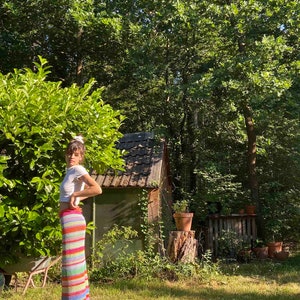K&ouml;nnte beinhalten: Eine Frau tr&auml;gt ein wei&szlig;es Crop-Top und einen bunten, gestreiften H&auml;kelrock und steht in einem grasbewachsenen Bereich mit einem kleinen Holzschuppen im Hintergrund.