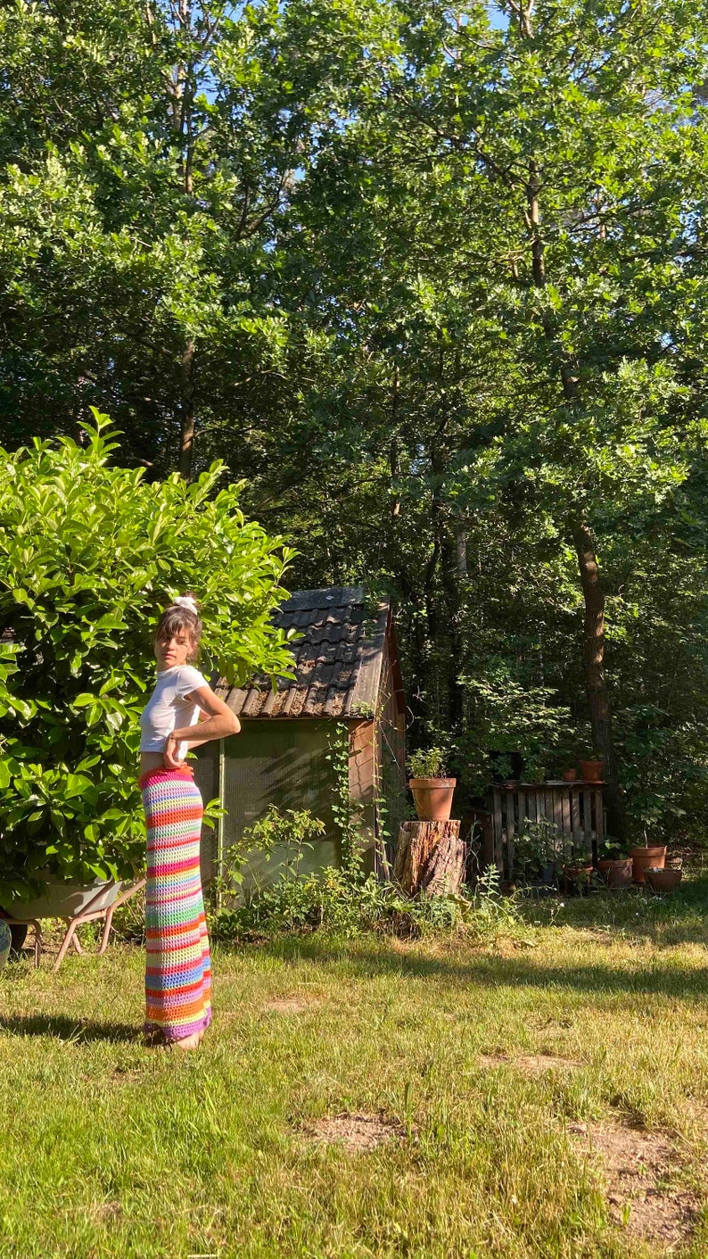K&ouml;nnte beinhalten: Eine Frau tr&auml;gt ein wei&szlig;es Crop-Top und einen bunten, gestreiften H&auml;kelrock und steht in einem grasbewachsenen Bereich mit einem kleinen Holzschuppen im Hintergrund.