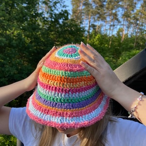 May include: A colorful crocheted bucket hat with stripes of pink, orange, green, purple, and white. The hat has a round, flat crown and a wide brim.